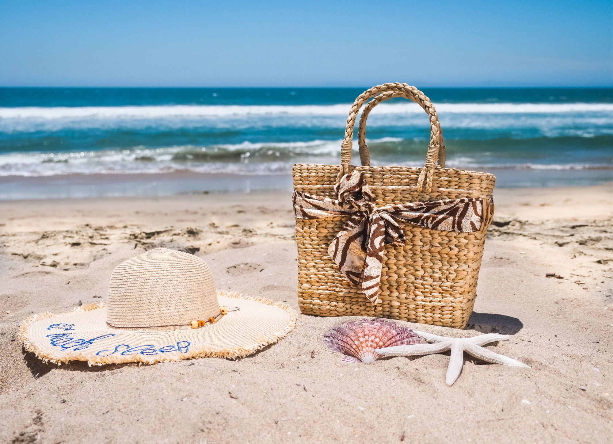 Panier en osier et chapeau de plage sur le sable, mettant en avant l'élégance, le respect de la nature et l'authenticité, valeurs de la boutique Mademoiselle K.
