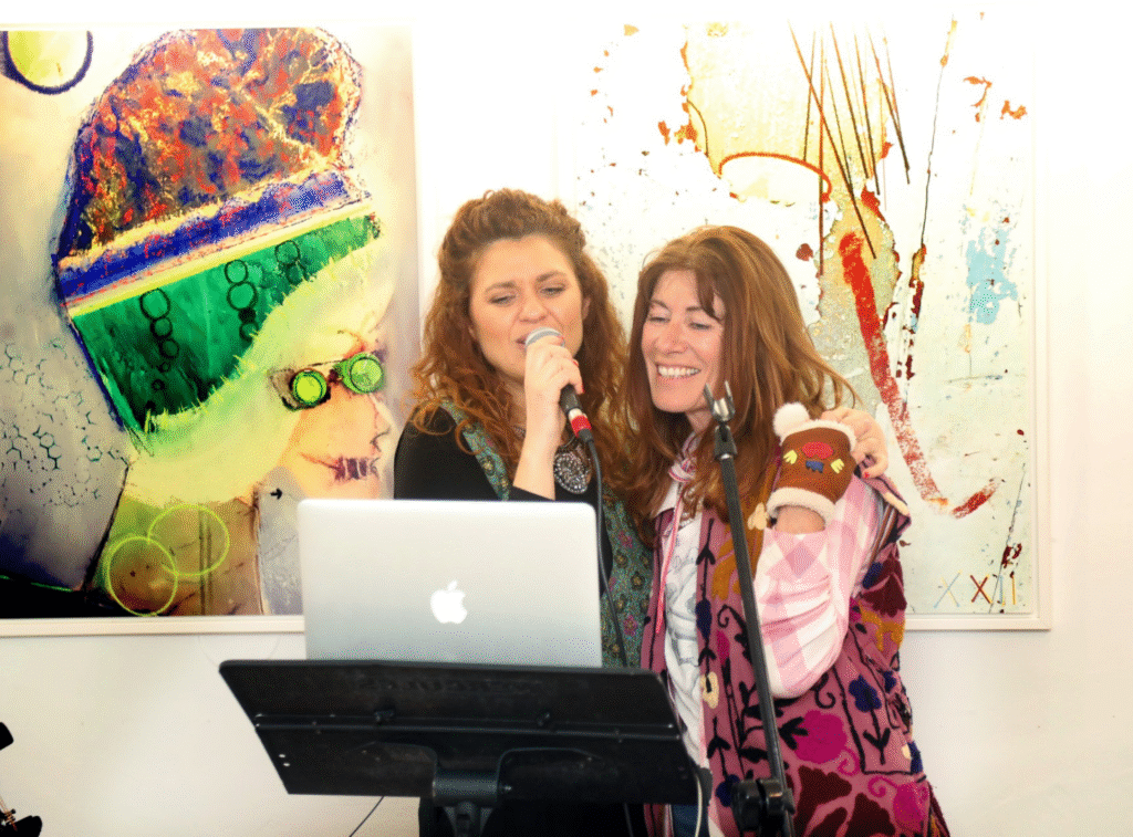 Photo de deux participantes chantant ensemble lors de l'inauguration de Mademoiselle K à Cassis, avec des œuvres d'art en arrière-plan, créant une ambiance festive et chaleureuse.