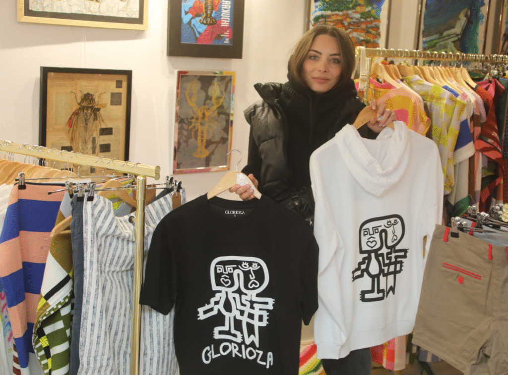 Une participante tient des t-shirts et des hoodies de la marque Glorioza lors de l'inauguration de Mademoiselle K à Cassis, entourée de vêtements colorés et d'œuvres d'art.